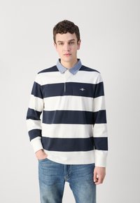 Tamsiai mėlynas ir baltas dryžuotas ilgomis rankovėmis polo marškinėlis su šviesiai džinsiniu apykakliu ir siuvinėta logotipu. Turi aptemptas rankogalius ir laisvą kirpimą.