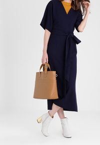 Marineblauwe jurk met een strik taille, korte mouwen en een V-hals; gecombineerd met een tan handtas en witte enkellaarsjes met gouden accenten.