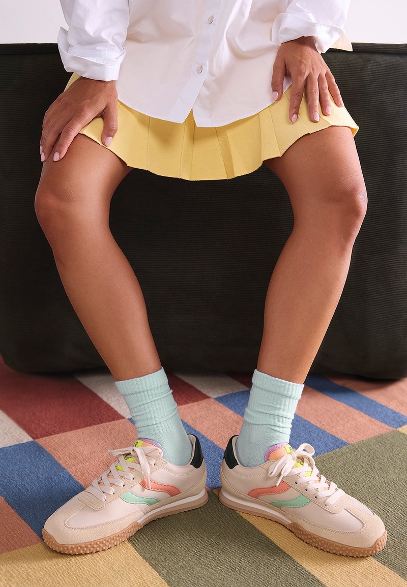 Jambes portant des baskets pastel et des chaussettes bleu clair, assises sur un canapé noir, vêtues d'une jupe jaune et d'une chemise blanche.