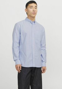 Camicia azzurra chiara a maniche lunghe con sottili righe verticali bianche e arancioni, colletto con bottoni e taschino sul petto. Indossata con jeans scuri.