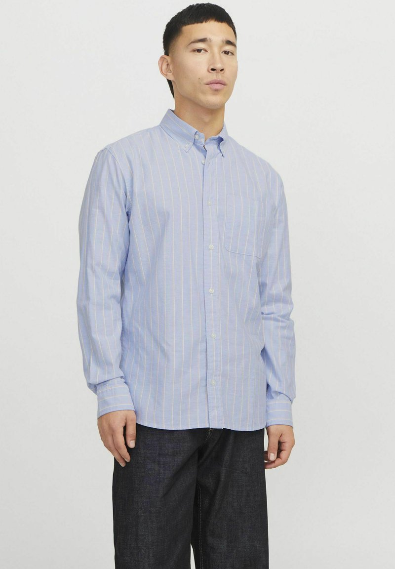 Camicia azzurra chiara a maniche lunghe con sottili righe verticali bianche e arancioni, colletto con bottoni e taschino sul petto. Indossata con jeans scuri.