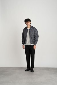 Grijze bomberjack met een gewatteerde textuur, geribde manchetten en een zijzak met rits, gecombineerd met een lichtgrijs sweater en zwarte broek.