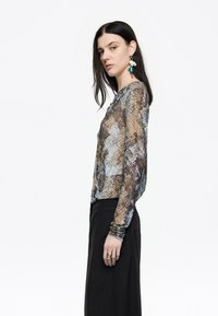 Vrouw met lang, donker haar die een doorzichtige, slangenprint long-sleeve top, zwarte broek, grote statement oorbellen en ringen draagt, staand in profiel.