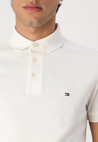 Polo shirt blanc en tissu texturé avec une patte de boutonnage à trois boutons et des manches courtes. Présente un petit logo bleu marine, rouge et blanc sur la poitrine.
