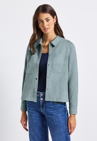 Žena s dlouhými hnědými vlasy má na sobě světlý tyrkysový kabát s knoflíky přes námořnický top a modré džíny, stojí na pozadí čistého bílého pozadí.