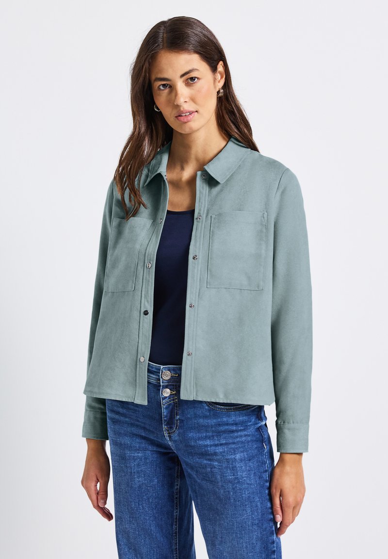 Žena s dlouhými hnědými vlasy má na sobě světlý tyrkysový kabát s knoflíky přes námořnický top a modré džíny, stojí na pozadí čistého bílého pozadí.