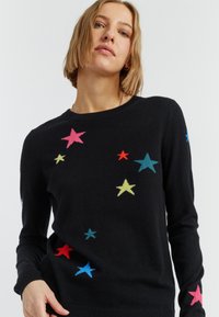 Zwarte trui met gekleurde sterrenpatronen in roze, blauw, groen en rood. Heeft een ronde halslijn en lange mouwen in een gladde textuur.