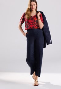 Red patterned blouse with short sleeves, paired with high-waisted, dark navy trousers and gold strappy sandals. The model holds a navy jacket.