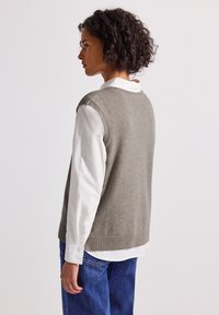 Kvinna med lockigt hår som bär en grå ärmlös tröja över en vit långärmad skjorta och blå jeans, vänd åt sidan mot en neutral bakgrund.