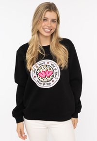Schwarzer Sweatshirt mit einem zentralen Grafikdesign, das ein Leopardenmuster und pinke Herzen zeigt, umgeben von dem Text "wild at heart" in einem kreisförmigen Design.