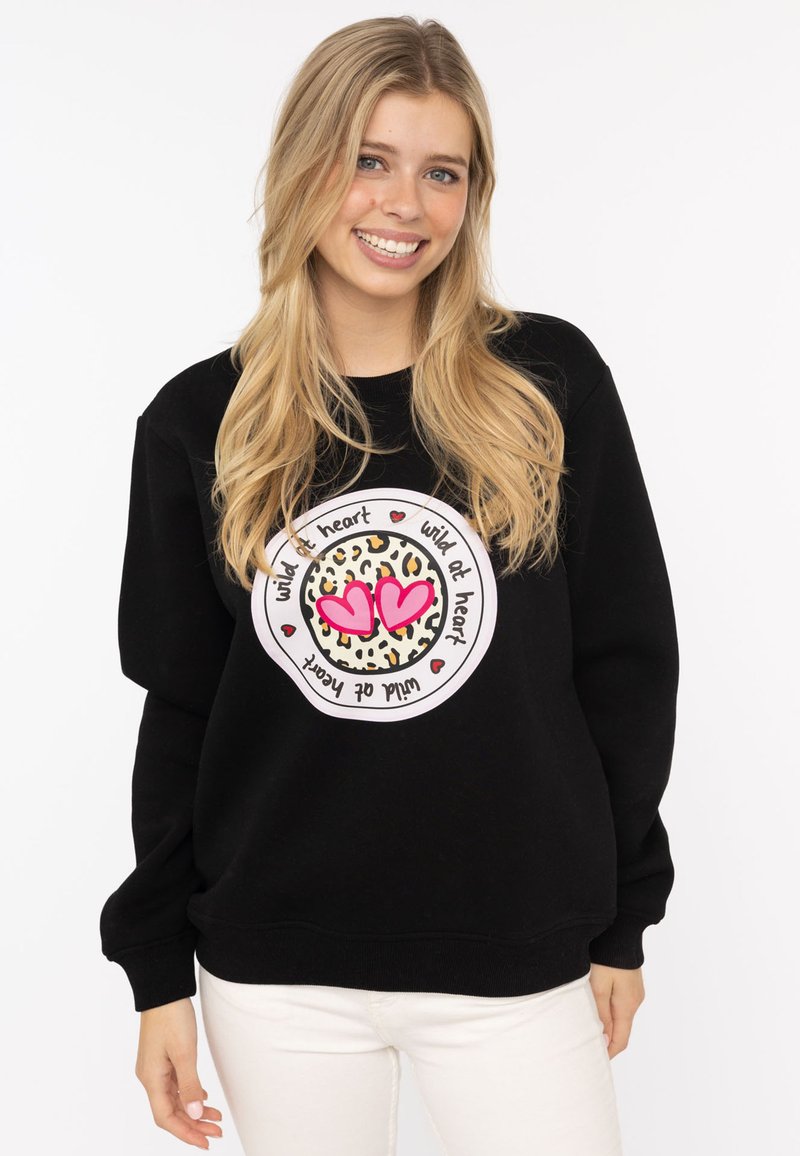 Schwarzer Sweatshirt mit einem zentralen Grafikdesign, das ein Leopardenmuster und pinke Herzen zeigt, umgeben von dem Text "wild at heart" in einem kreisförmigen Design.