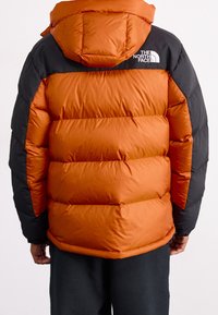 Doudoune orange et noire avec capuche, comportant des sections matelassées et le logo The North Face en blanc dans le haut du dos.