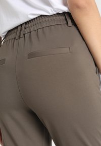 Elastische taillebandbroek in donkerbruine stof met een gladde textuur, voorzien van twee achterzakken en subtiele zijdetails.