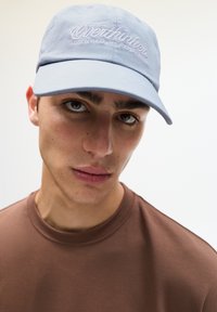 Light blue cap with embroidered text in white, set against a model wearing a brown t-shirt. Focus on cap's soft fabric and structured shape.