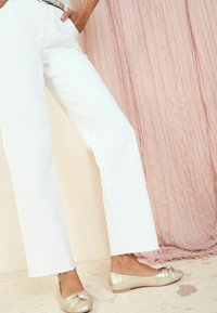 Witte wijde jeans met een rafelige zoom, gecombineerd met metallic gouden ballerina's met een knoopdetail op de teen. De achtergrond is van gestructureerde roze stof.