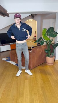 Temno modra zip-up kapuca z logotipom na levem prsnem delu, sivi oprijeti džins, in bele tene, stoji na lesenem tleh.