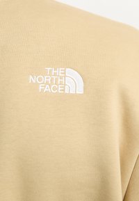 Beige mikina s vyšitým bílým logem "The North Face" na horním levém rohu. Měkký materiál a uvolněný střih.