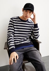 Navy- und grau gestreifter Pullover mit Rundhalsausschnitt, über einer blauen Schicht getragen, kombiniert mit dunkelgrauen Jeans und einer schwarzen Kapuze.