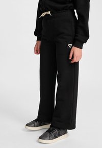 Schwarze Fleece-Sweatpants mit einem Kordelzugbund, weitem Bein und einem kleinen Logoakzent. Kombiniert mit glitzernden schwarzen Turnschuhen.