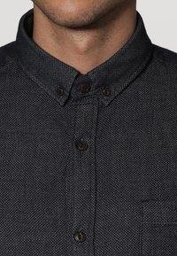 Camicia blu scuro con bottoni, realizzata in tessuto testurizzato, con colletto a punta, bottoni marroni e taschino sul lato sinistro.