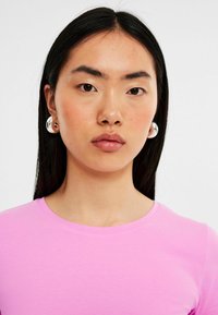 Roze fitted T-shirt met een ronde halslijn; model draagt metallic zilveren ringoorbellen. Glad stof, natuurlijk haar valt recht.