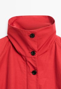 Veste rouge à col haut, dotée de boutons noirs et d'un tissu lisse et texturé. Le design met en avant des lignes épurées et une silhouette structurée.