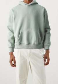 Sudadera verde menta hecha de un tejido suave, con un bolsillo frontal, puños de canalé y capucha con cordón. Combinada con pantalones blancos.