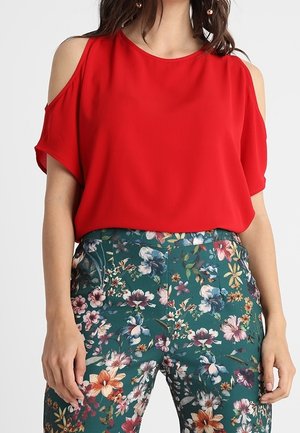 Kobieta ubrana w czerwoną bluzkę z odkrytymi ramionami, w zestawieniu z zielonymi spodniami w kwiatowy wzór, stojąca na jednolitym tle.