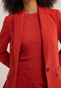 Červený měkký blazer s ostrými klopami, kombinovaný s červeným žebrovaným topem, který má přiléhavý střih a jemnou strukturu. Viditelný detail s černým knoflíkem.