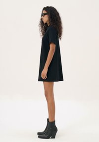 Vrouw met lang, krullend haar, die zwarte zonnebril, een korte zwarte jurk en zwarte hoge enkelboots draagt, staand in profiel tegen een witte achtergrond.