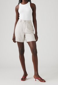 Nainen, jolla on yllään valkoinen hihaton paita, beigekirjailut korkeat shortsit ja punaiset korkokengät, seisoo vaaleaa taustaa vasten.