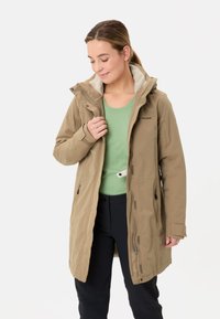 Donna che indossa un cappotto beige con cappuccio sopra una maglietta verde e pantaloni neri, mentre tiene la cerniera del cappotto, guardando in basso e sorridendo leggermente.