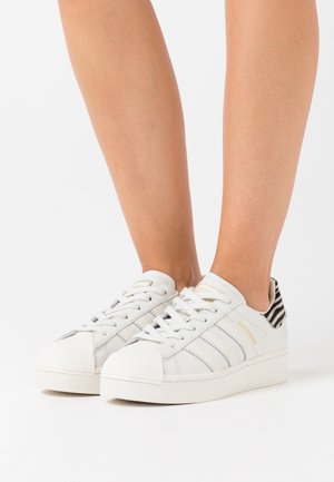 Bílé plátěné tenisky se šedými pruhovanými detaily a strukturovanou špičkou, s dekorovaným podpatkem a rovnými tkaničkami.