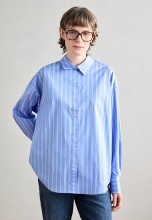Blouse bleu clair à manches longues avec des rayures verticales blanches, fermeture à boutons sur le devant et col classique. Portée par-dessus un pantalon en denim, coupe décontractée.