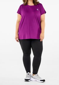 Kvinde iført en løs lilla t-shirt, sorte leggins og hvide løbesko står foran en hvid baggrund.