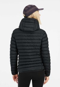 Zwarte pufferjack met horizontale quiltmotieven, een getailleerde capuchon en lange mouwen. Gecombineerd met een donkere pet met een gestreepte textuur.