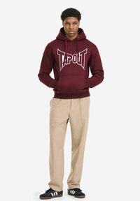 Mladić stoji, nosi tamnocrvenu majicu s kapuljačom s natpisom "TAPOUT", bež hlače i crne tenisice s bijelim prugama, ruke u džepovima kapuljače.