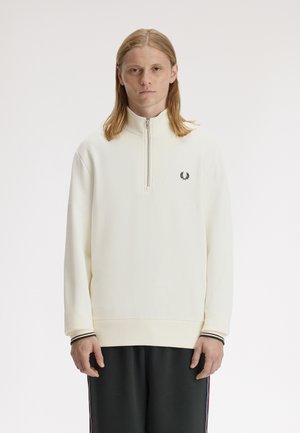 Jeune homme aux cheveux longs portant un sweat-shirt blanc à fermeture éclair quart de zip avec un petit logo de couronne de laurier noir sur la poitrine, associé à un pantalon noir.