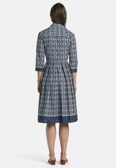 Frau mit langen braunen Haaren, die ein knieumspielendes blau-weiß gemustertes Kleid mit dreiviertel Ärmeln trägt, steht mit dem Rücken zum Betrachter vor einem weißen Hintergrund.