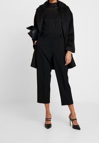 Schwarzes, gemustertes Spitzentop unter einem schwarzen, strukturierten Mantel, kombiniert mit schwarzen, cropped Hosen und schwarzen, spitzen Pumps.