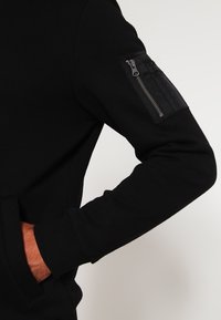 Felpa nera con tasca con zip sulla manica sinistra, caratterizzata da una texture liscia e polsini aderenti. Tasca a marsupio sul davanti.