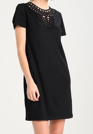 Woman wearing a short-sleeve black dress with a decorative cutout lace pattern around the neckline.