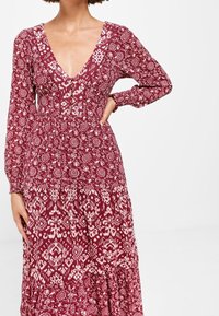 Langärmliges Kleid mit tiefem V-Ausschnitt in einem rot-weißen Blumenmuster, ausgestattet mit einem taillierten Oberteil und einem schichtartigen, fließenden Rock mit Knopfdetails.