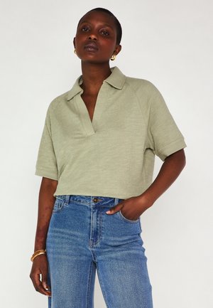 Femme portant un polo à manches courtes vert clair et un jean taille haute bleu, avec une main dans la poche, des boucles d'oreilles et des bracelets en or.