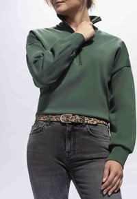 Donna che indossa una felpa verde con cerniera a quarto di zip, jeans grigio scuro e una cintura leopardata, che si sistema il colletto con la mano destra.