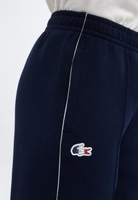 Laivastonsiniset collegehousut, joissa valkoinen reunus sivusaumassa ja pieni kirjailtu krokotiililogo punaisena, valkoisena ja sinisenä lähellä taskua.