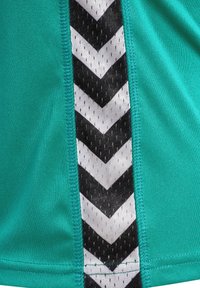 Top de tela verde azulado con un detalle en chevron en negro y blanco a lo largo del costado, que presenta un patrón texturizado y secciones con aspecto de malla.