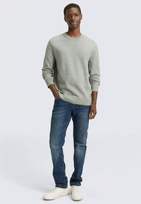 Hellgrauer Strickpullover mit Rundhalsausschnitt, kombiniert mit dunkelblauen Jeans und weißen Sneakers. Das Model steht entspannt.