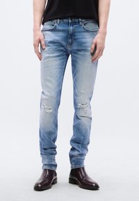 Ανοιχτό μπλε τζιν με φθαρμένες λεπτομέρειες, στενή εφαρμογή και κοντράστ ραφές. Φοριέται με σκούρα καφέ μποτάκια μέχρι τον αστράγαλο.