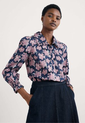 Vrouw in een marineblauw shirt met roze bloemenpatronen en een donkere denim rok, staand met de handen in de zakken tegen een effen achtergrond.